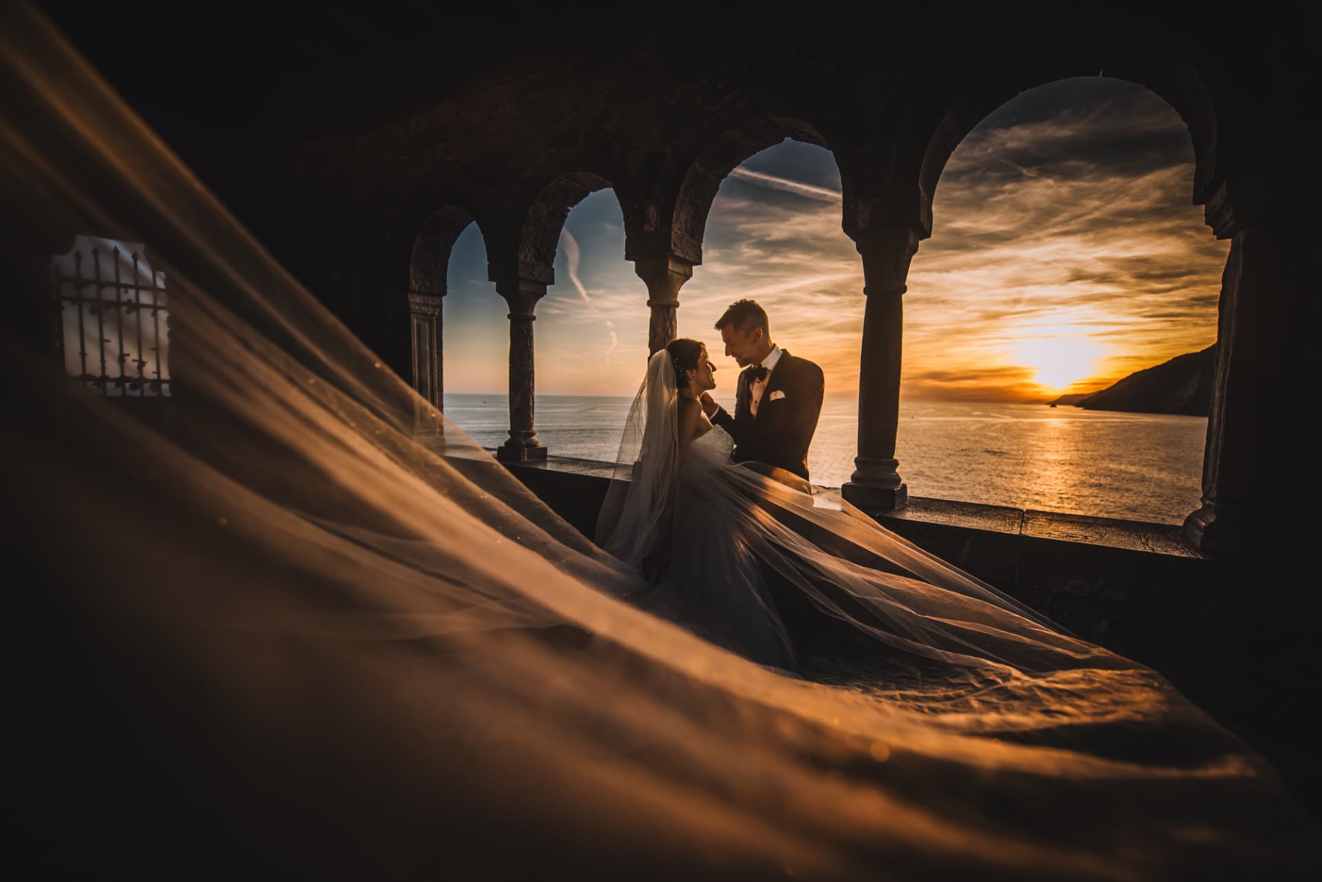 Gli sposi sotto gli archi di Portovenere con il velo che attraversa la scena al tramonto