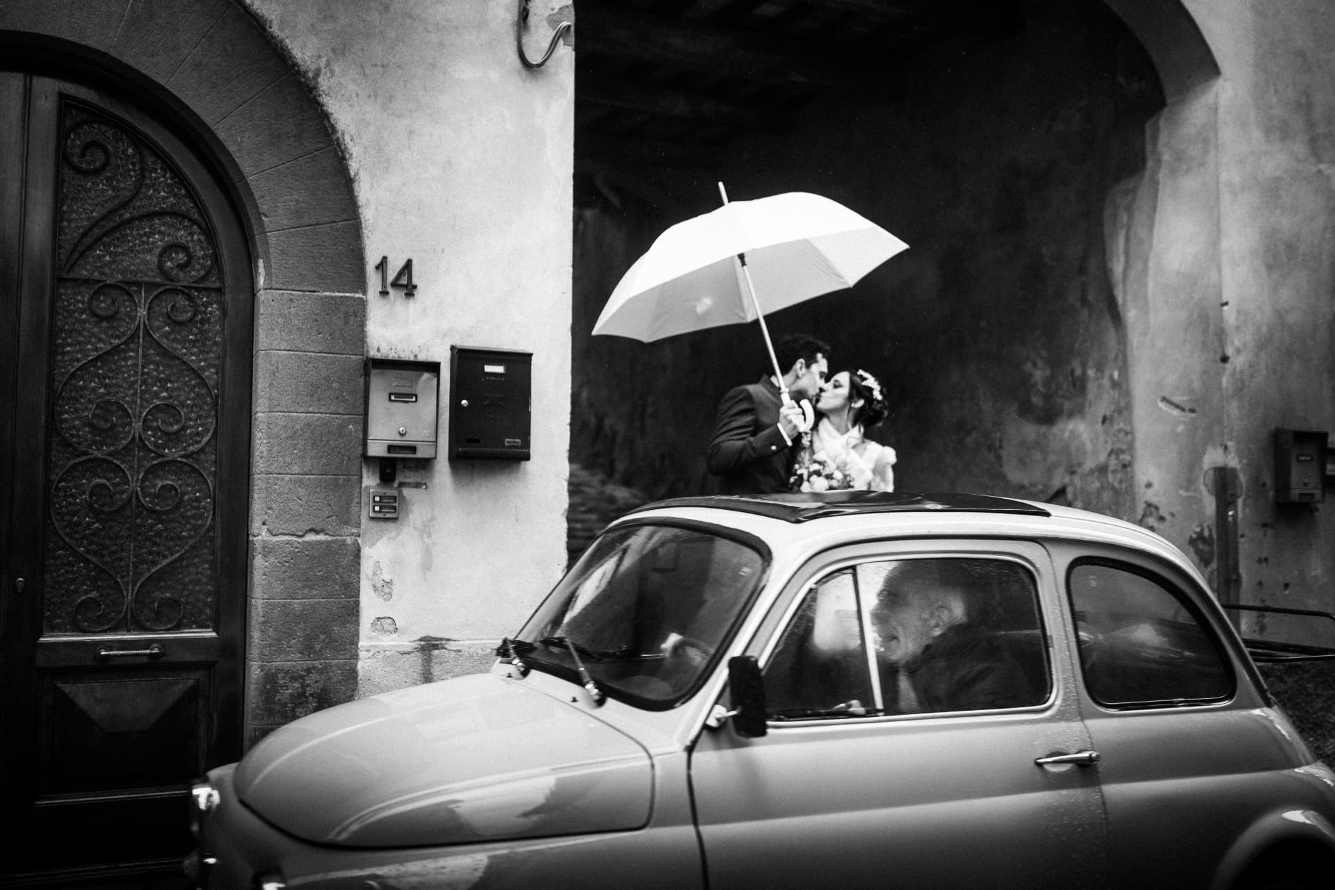 Bacio degli sposi sotto l'ombrello, oltre una vecchia auto nel vicolo